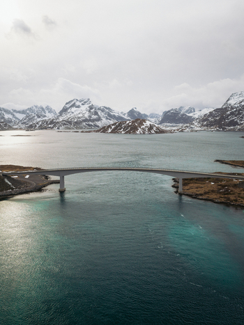Aerial view of a bridge spanning turquoise waters framed by snow dusted peaks and rugged shores Flakstadbruene Nordland Norway. Aerial view of a bridge spanning turquoise waters framed by snow dusted peaks and rugged shores Flakstadbruene Nordland Norway.