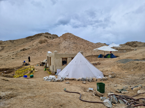 View of a white conical tent and a small mud brick building in a rugged desert landscape with mountains under a cloudy sky in Leh Ladakh India.