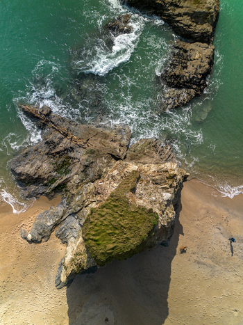 Aerial view of a large rock formation on the sandy beach with turquoise water crashing waves and a person walking with a dog in Carlyon Bay Cornwall United Kingdom. Aerial view of a large rock formation on the sandy beach with turquoise water crashing waves and a person walking with a dog in Carlyon Bay Cornwall United Kingdom.
