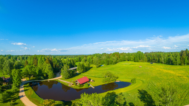 Aerial view of a small house with a red roof situated beside a peaceful pond in a lush green rural landscape with trees and meadows in LATVIA. Aerial view of a small house with a red roof situated beside a peaceful pond in a lush green rural landscape with trees and meadows in LATVIA.