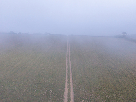 Aerial view of misty rural countryside with a dirt path cutting through agricultural farmland under a thick layer of fog in Looe Cornwall United Kingdom. Aerial view of misty rural countryside with a dirt path cutting through agricultural farmland under a thick layer of fog in Looe Cornwall United Kingdom.