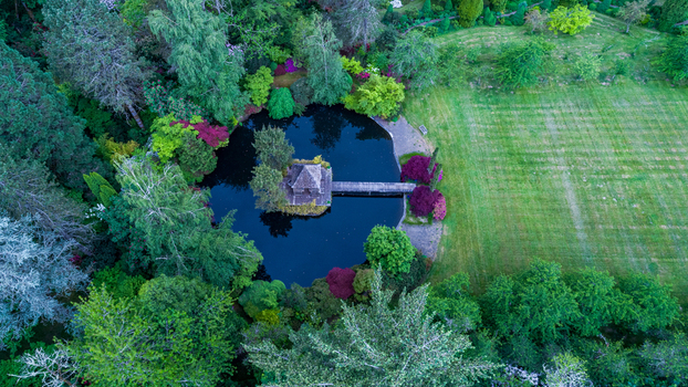 Aerial view of a serene garden pond featuring a small gazebo on an island connected by a wooden bridge surrounded by lush green trees and colorful shrubs in Portland Oregon United States. Aerial view of a serene garden pond featuring a small gazebo on an island connected by a wooden bridge surrounded by lush green trees and colorful shrubs in Portland Oregon United States.