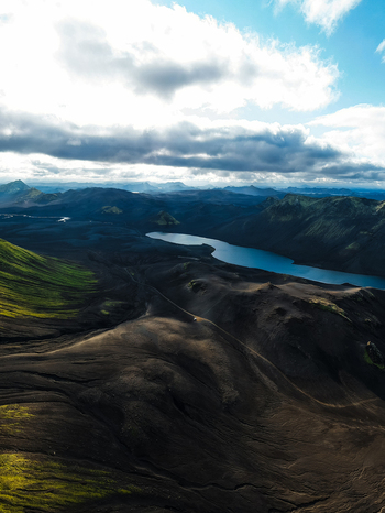 Aerial view of stark contrasting landscapes where dark volcanic earth meets vibrant green moss and a serene blue lake reflects the sky Landmannalaugar Iceland. Aerial view of stark contrasting landscapes where dark volcanic earth meets vibrant green moss and a serene blue lake reflects the sky Landmannalaugar Iceland.