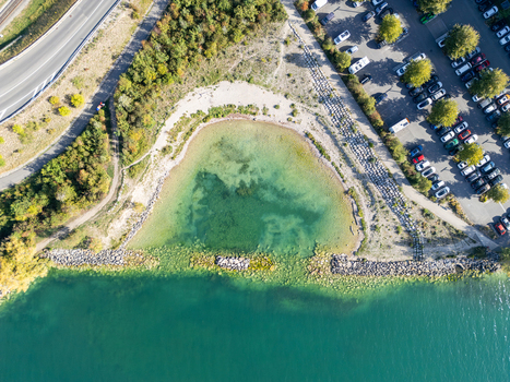 Aerial view of a small bay with turquoise water rocky shoreline adjacent parking lot and surrounding greenery in Neuenburg Neuenburg Switzerland.