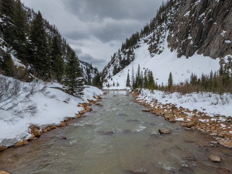 View of a flowing river through a snowy mountain valley with evergreen trees and rocky slopes under an overcast sky Colorado United States.