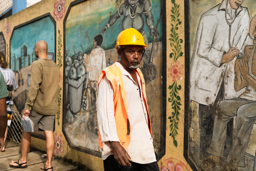 Sri Lanka 20 February 2026 View of a worker wearing a yellow hard hat and orange safety vest walking past a wall decorated with colorful murals.