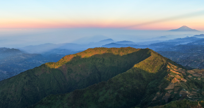Aerial view of Mount Bismo illuminated by golden hour light with lush green slopes and distant volcanic peaks in Kecamatan Kejajar Jawa Tengah Indonesia.