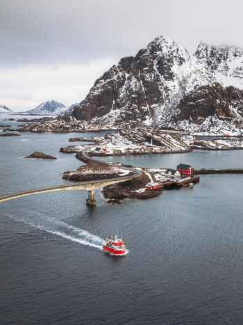 Aerial view of a vibrant red boat cutting through the dark waters towards the cluster of buildings at Henningsv&aelig;r Nordland Norway.