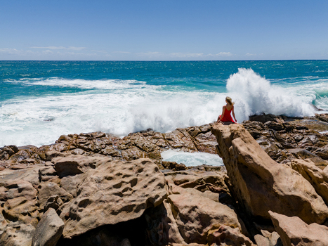 Aerial view of Injidup Natural Spa with a woman in a red dress sitting on the rugged coastal rocks as waves crash against the shore in Yallingup Western Australia Australia. Aerial view of Injidup Natural Spa with a woman in a red dress sitting on the rugged coastal rocks as waves crash against the shore in Yallingup Western Australia Australia.