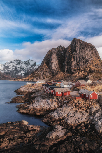 View of traditional red fishing huts on the rocky shore of Hamnoy with a steep mountain peak in the background Hamnoy Nordland Norway.