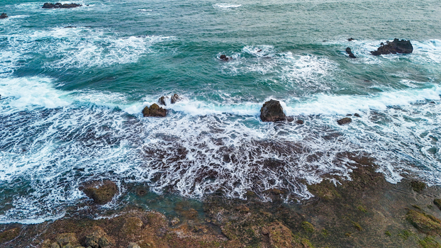 Aerial view of waves crashing against rocky coastline with mossy stones and turquoise water in Indonesia. Aerial view of waves crashing against rocky coastline with mossy stones and turquoise water in Indonesia.