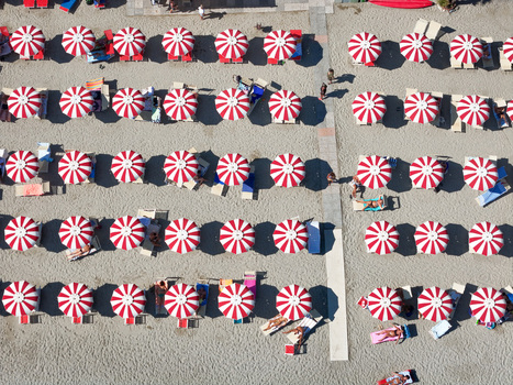 Aerial view of rows of red and white striped beach umbrellas on the sandy beach with people relaxing on lounge chairs in Bergeggi Liguria Italy. Aerial view of rows of red and white striped beach umbrellas on the sandy beach with people relaxing on lounge chairs in Bergeggi Liguria Italy.