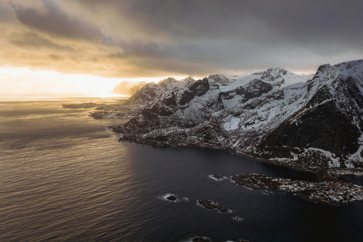 Aerial view of snow dusted mountains meet the dark rippling waters under a dramatic sky where the sun casts a golden glow Reinebringen Nordland Norway.