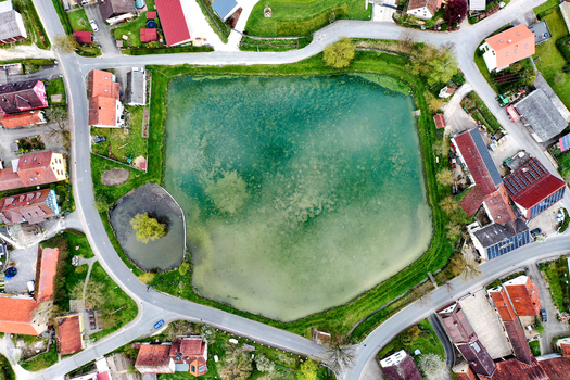 Aerial view of the Ailsbach pond surrounded by traditional houses with red roofs and winding roads in a rural village Lonnerstadt Bayern Germany. Aerial view of the Ailsbach pond surrounded by traditional houses with red roofs and winding roads in a rural village Lonnerstadt Bayern Germany.