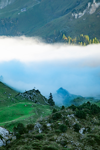 Aerial view of the Swiss Alps in Airolo featuring a small mountain chalet nestled among green slopes and rocky outcrops partially obscured by low hanging clouds in Airolo Ticino Switzerland. Aerial view of the Swiss Alps in Airolo featuring a small mountain chalet nestled among green slopes and rocky outcrops partially obscured by low hanging clouds in Airolo Ticino Switzerland.