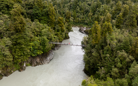 Aerial view of a narrow suspension bridge stretching over the milky glacial river embraced by lush green rainforest Kokatahi West Coast Region New Zealand.