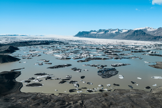 Aerial view of a glacial lagoon filled with floating icebergs surrounded by dark volcanic terrain and distant snow capped mountains in Sveitarfelagid Hornafjordur Iceland. Aerial view of a glacial lagoon filled with floating icebergs surrounded by dark volcanic terrain and distant snow capped mountains in Sveitarfelagid Hornafjordur Iceland.