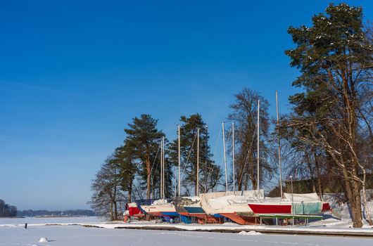 View of sailboats stored on the snow covered shore of a frozen lake surrounded by tall pine trees under a clear blue sky in Trakai Vilnius County Lithuania. View of sailboats stored on the snow covered shore of a frozen lake surrounded by tall pine trees under a clear blue sky in Trakai Vilnius County Lithuania.