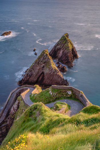 View of the winding stone path of Dunquin Pier leading to the Atlantic Ocean with sharp sea stacks and grassy cliffs Dun Chaoin County Kerry Ireland. View of the winding stone path of Dunquin Pier leading to the Atlantic Ocean with sharp sea stacks and grassy cliffs Dun Chaoin County Kerry Ireland.