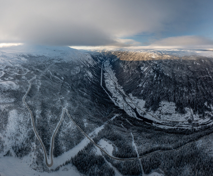 Aerial view of the winding mountain road leading through the snow covered valley near Rjukan surrounded by dramatic peaks and evergreen forests in Rjukan Telemark Norway. Aerial view of the winding mountain road leading through the snow covered valley near Rjukan surrounded by dramatic peaks and evergreen forests in Rjukan Telemark Norway.