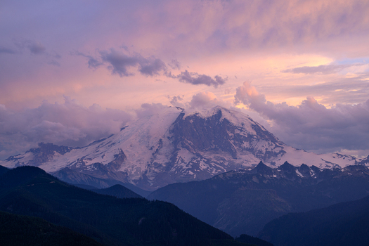 View of Mount Rainier at sunset with snow capped peaks purple and pink clouds and dark forested mountains in the foreground in Pierce County Washington United States. View of Mount Rainier at sunset with snow capped peaks purple and pink clouds and dark forested mountains in the foreground in Pierce County Washington United States.