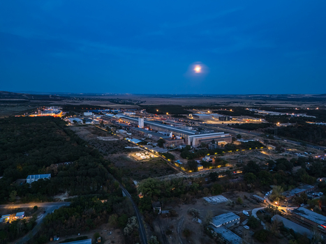 Aerial view of an illuminated aluminum smelter and industrial complex beside rail lines amid woodland and farmland under a moonlit evening sky in Inota Hungary.