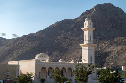 View of a mosque with a tall minaret and domes set against a rugged mountain landscape in Muscat Muscat Governorate Oman. View of a mosque with a tall minaret and domes set against a rugged mountain landscape in Muscat Muscat Governorate Oman.