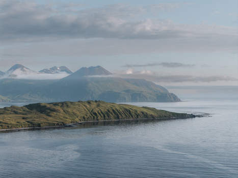 Aerial view of Captains Bay featuring lush green hills calm coastal waters and distant mountains partially obscured by low hanging clouds in Unalaska Alaska United States. Aerial view of Captains Bay featuring lush green hills calm coastal waters and distant mountains partially obscured by low hanging clouds in Unalaska Alaska United States.