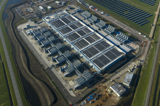 Aerial view of an AI data center and cloud computing hub exterior featuring rooftop cooling towers and solar panels surrounded by green fields. Aerial view of an AI data center and cloud computing hub exterior featuring rooftop cooling towers and solar panels surrounded by green fields.