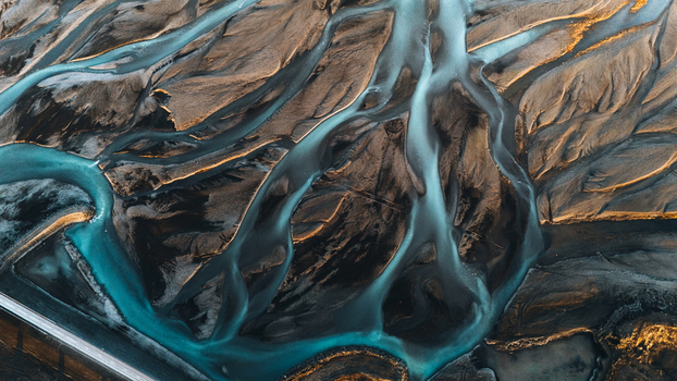 Aerial view of intricate braided glacial river channels flowing through dark volcanic sediment with soft golden light highlights Skaftarhreppur Iceland.