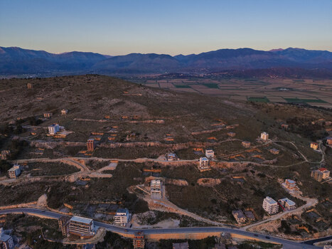 Aerial view of the hillside landscape featuring scattered residential buildings winding roads and distant mountains under the warm light of golden hour in Saranda Vlore County Albania. Aerial view of the hillside landscape featuring scattered residential buildings winding roads and distant mountains under the warm light of golden hour in Saranda Vlore County Albania.