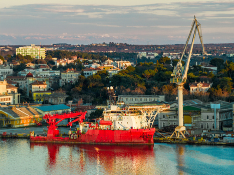 Aerial view of the Uljanik Shipyard in Pula featuring a large red vessel docked near a tall industrial crane with the city skyline and golden hour light in the background Pula Croatia. Aerial view of the Uljanik Shipyard in Pula featuring a large red vessel docked near a tall industrial crane with the city skyline and golden hour light in the background Pula Croatia.
