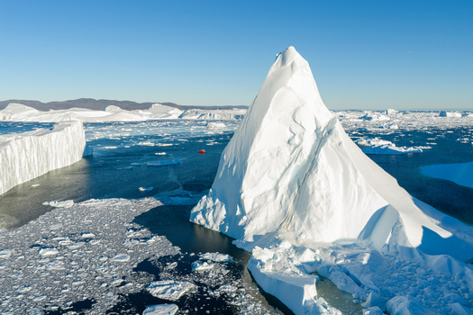 Aerial view of the Ilulissat glacier with a massive iceberg floating in the deep blue sea surrounded by smaller ice fragments and a clear sky in Greenland. Aerial view of the Ilulissat glacier with a massive iceberg floating in the deep blue sea surrounded by smaller ice fragments and a clear sky in Greenland.