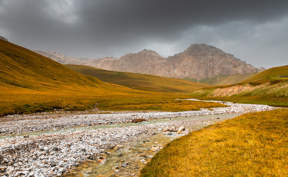 View of the rocky riverbed of Kol Suu flowing through golden grassy hills under dramatic storm clouds with rugged mountains in the background in At Bashi District Naryn Region Kyrgyzstan. View of the rocky riverbed of Kol Suu flowing through golden grassy hills under dramatic storm clouds with rugged mountains in the background in At Bashi District Naryn Region Kyrgyzstan.