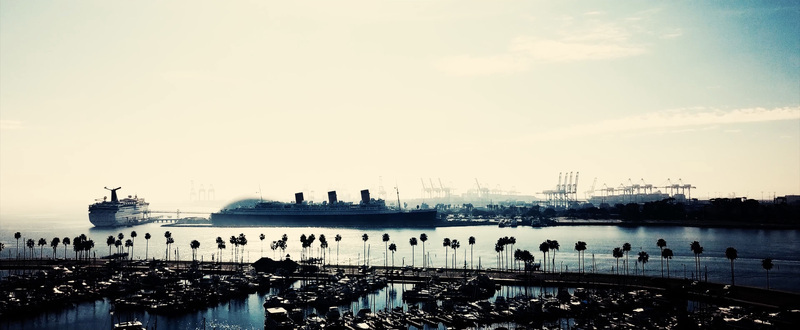 Aerial view of the historic RMS Queen Mary docked in the harbor with a cruise ship palm trees and industrial cranes in Long Beach California United States.