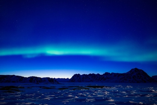 Aerial view of the Aurora Borealis glowing green over snow capped mountain peaks and an icy landscape under a starry night sky in Tasiilaq Sermersooq Municipality Greenland. Aerial view of the Aurora Borealis glowing green over snow capped mountain peaks and an icy landscape under a starry night sky in Tasiilaq Sermersooq Municipality Greenland.