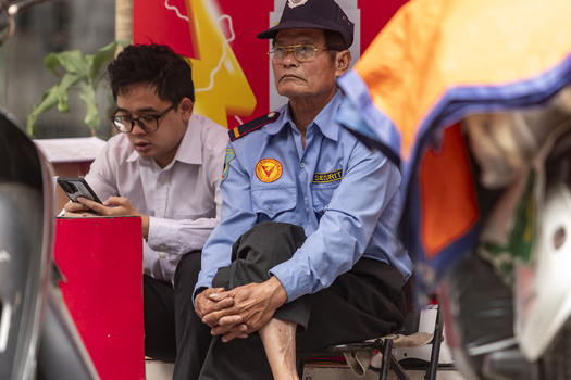 Hanoi Ha Noi Vietnam 11 November 2025 View of a security guard in a blue uniform sitting next to a man looking at his smartphone in an urban setting.