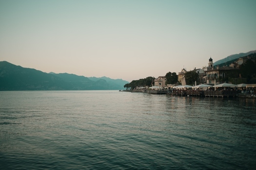 View of Lake Garda with historic buildings and restaurant terraces along the waterfront under a clear sky and distant mountains Italy. View of Lake Garda with historic buildings and restaurant terraces along the waterfront under a clear sky and distant mountains Italy.
