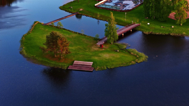Aerial view of a small grassy island with trees and a wooden footbridge in a calm lake in Latvia. Aerial view of a small grassy island with trees and a wooden footbridge in a calm lake in Latvia.