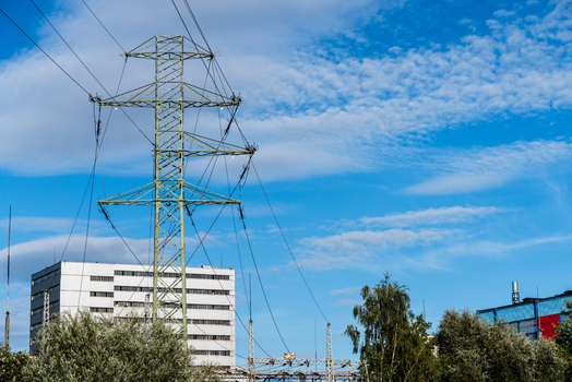 View of an electricity transmission tower with power lines against a blue sky with clouds and an industrial building in the background in Poland.
