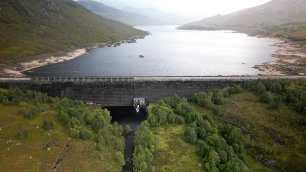 Aerial view of Cluanie Dam with its concrete structure holding back the reservoir surrounded by rolling hills and lush greenery in Inverness Scotland United Kingdom.