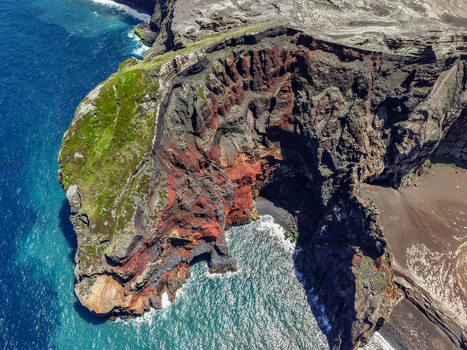 Aerial view of the rugged Capelinhos Volcano coastline where stark black volcanic rock meets vibrant green vegetation and turquoise waters Capelo A&ccedil;ores Portugal.