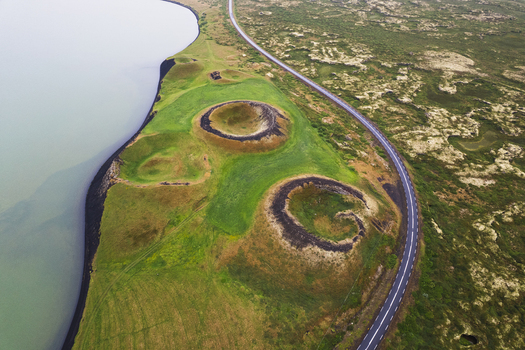 Aerial view of winding road around pseudocraters dotting the vibrant green landscape contrasting with the dark volcanic earth and the serene lake Lake Myvatn Iceland.