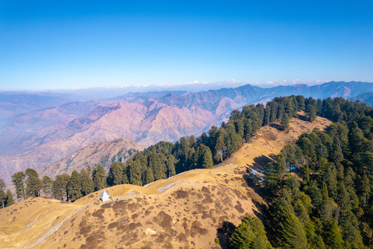Aerial view of the mountain ridges featuring a small white temple and dense pine forests under a clear blue sky in Chakrata Uttarakhand India. Aerial view of the mountain ridges featuring a small white temple and dense pine forests under a clear blue sky in Chakrata Uttarakhand India.