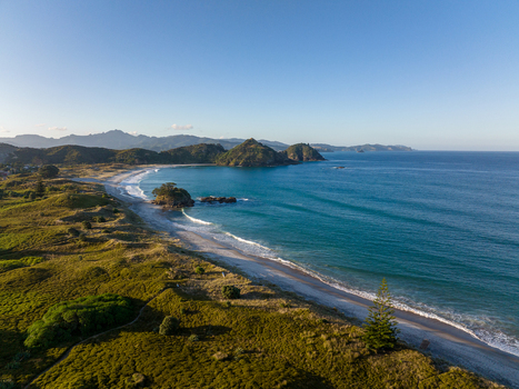 Aerial view of the coastline with rolling green hills sandy beach turquoise ocean waves and distant mountains under a clear blue sky in Great Barrier Island New Zealand. Aerial view of the coastline with rolling green hills sandy beach turquoise ocean waves and distant mountains under a clear blue sky in Great Barrier Island New Zealand.