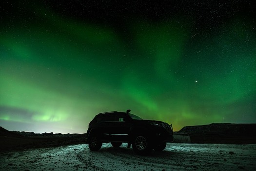 View of a dark SUV parked on a snowy terrain under the vibrant dancing green and white hues of the Aurora Borealis Krysuvikurkirkja Hafnarfjardarkaupstadur Iceland.