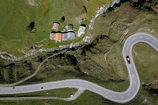 Aerial view of the winding Julier Pass road cutting through the green alpine landscape with small mountain houses and a stream in Surses Grisons Switzerland. Aerial view of the winding Julier Pass road cutting through the green alpine landscape with small mountain houses and a stream in Surses Grisons Switzerland.