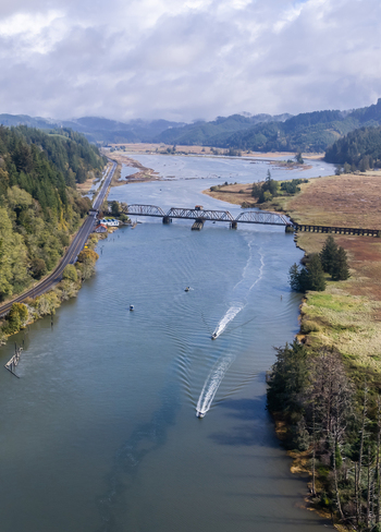 Aerial view of the Siuslaw River Train Bridge spanning the river with boats leaving wakes surrounded by lush green hills and a coastal highway in Florence Oregon United States.