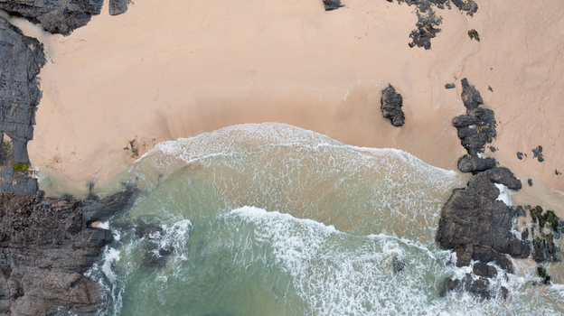 Aerial view of Harlyn Bay Beach featuring golden sand and turquoise waves crashing against dark jagged rocks in Saint Merryn Cornwall United Kingdom.