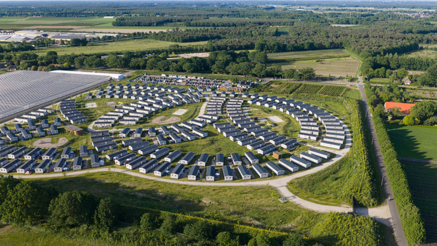 Aerial view of a holiday park featuring numerous uniform chalets arranged in curved rows amidst green fields and trees near a large greenhouse complex in Venlo Limburg Netherlands. Aerial view of a holiday park featuring numerous uniform chalets arranged in curved rows amidst green fields and trees near a large greenhouse complex in Venlo Limburg Netherlands.